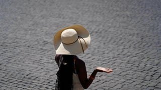 Eine Frau versucht sich mit einem Hut vor der Sonne zu Schützen. Sie ist auf einem Pflasterstein-Platz. Solche Plätze sind der Grund, warum viele Städte in Rheinland-Pfalz beim Hitze-Check der Deutschen Umwelthilfe schlecht abschneiden.