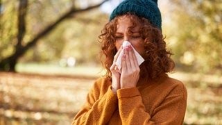 Eine Frau mit blauer Strickmütze und gelbem Pullover niest in ihr Taschentuch, weil sie eine Grippe oder Erkältung hat und sich davor nicht schützen konnte